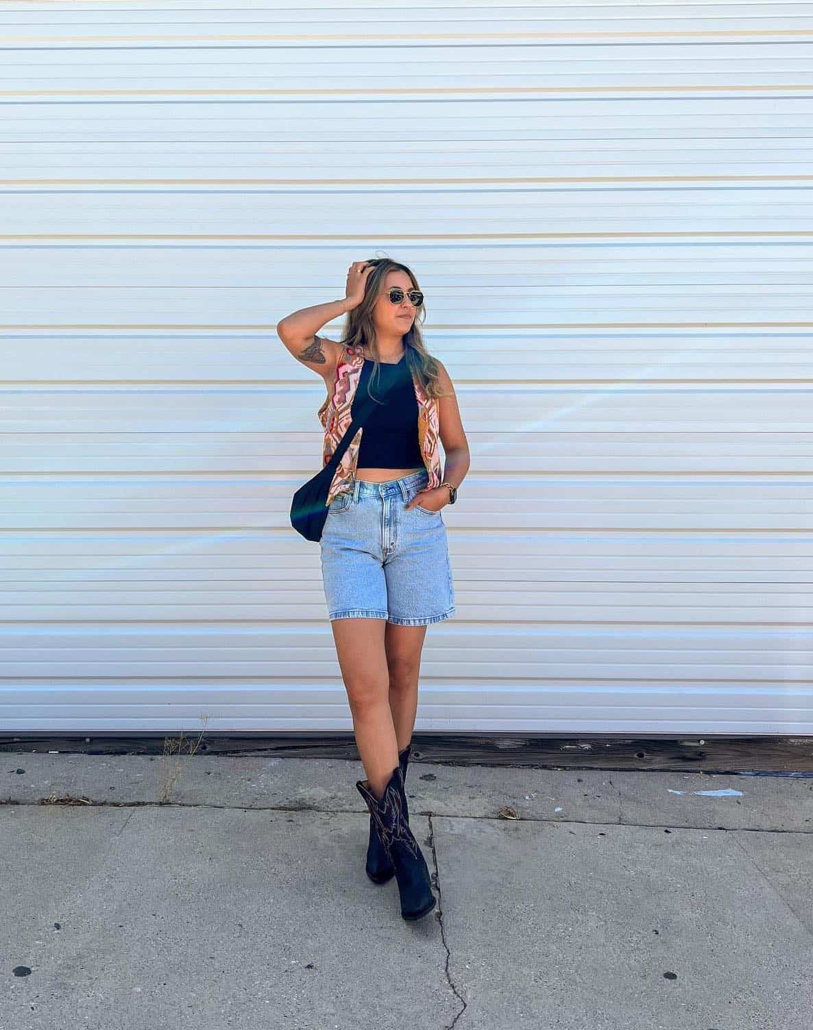 A woman wearing denim shorts with a black crop top, a colorful patterned vest, short black cowboy boots, and a black crossbody bag