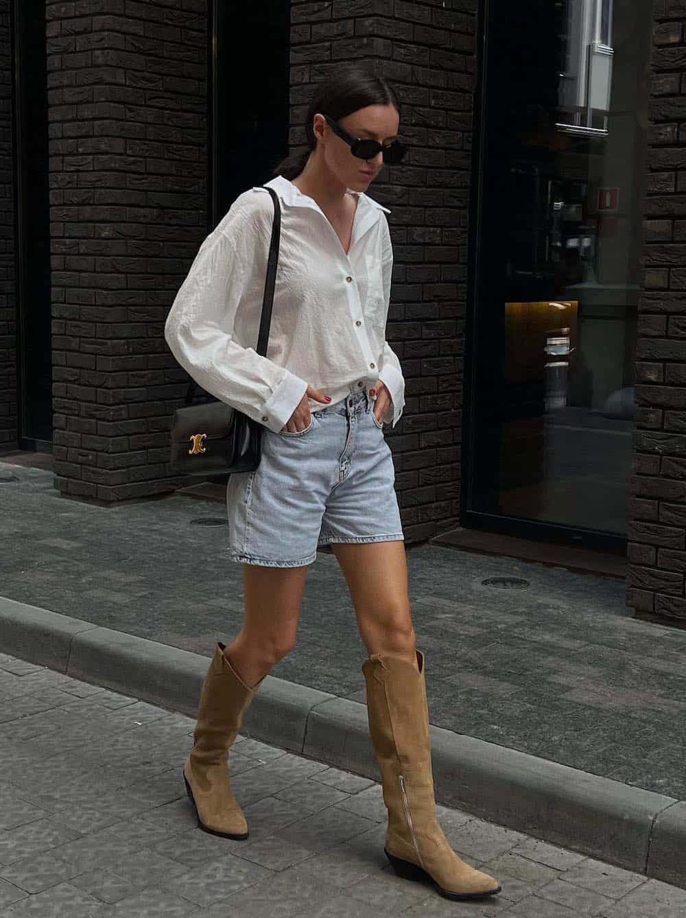 A woman wearing light-washed denim shorts with tan suede cowboy boots, a white button-up, and a black shoulder bag