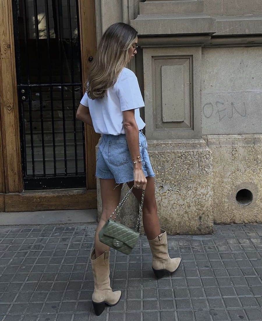 A woman wearing denim shorts with a white tee, short beige Western boots, and a green shoulder bag