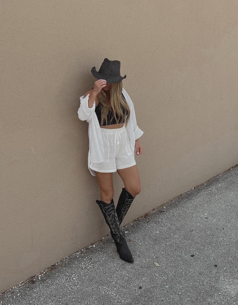 A woman wearing a matching white linen set with a button-up top and drawstring shorts with tall black cowboy boots, a black cropped tank, and a black cowboy hat