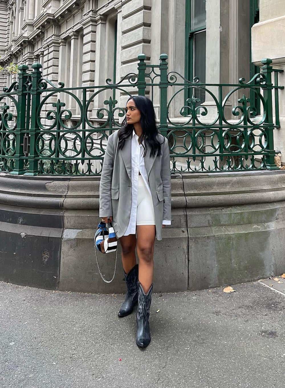 A woman wearing white running shorts with a white button-up, an oversized grey blazer, and short black Western-style boots