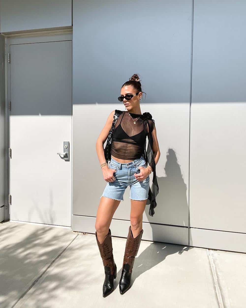 A woman wearing a black bralette, a black mesh top, light-washed denim shorts, and espresso brown and black cowboy boots