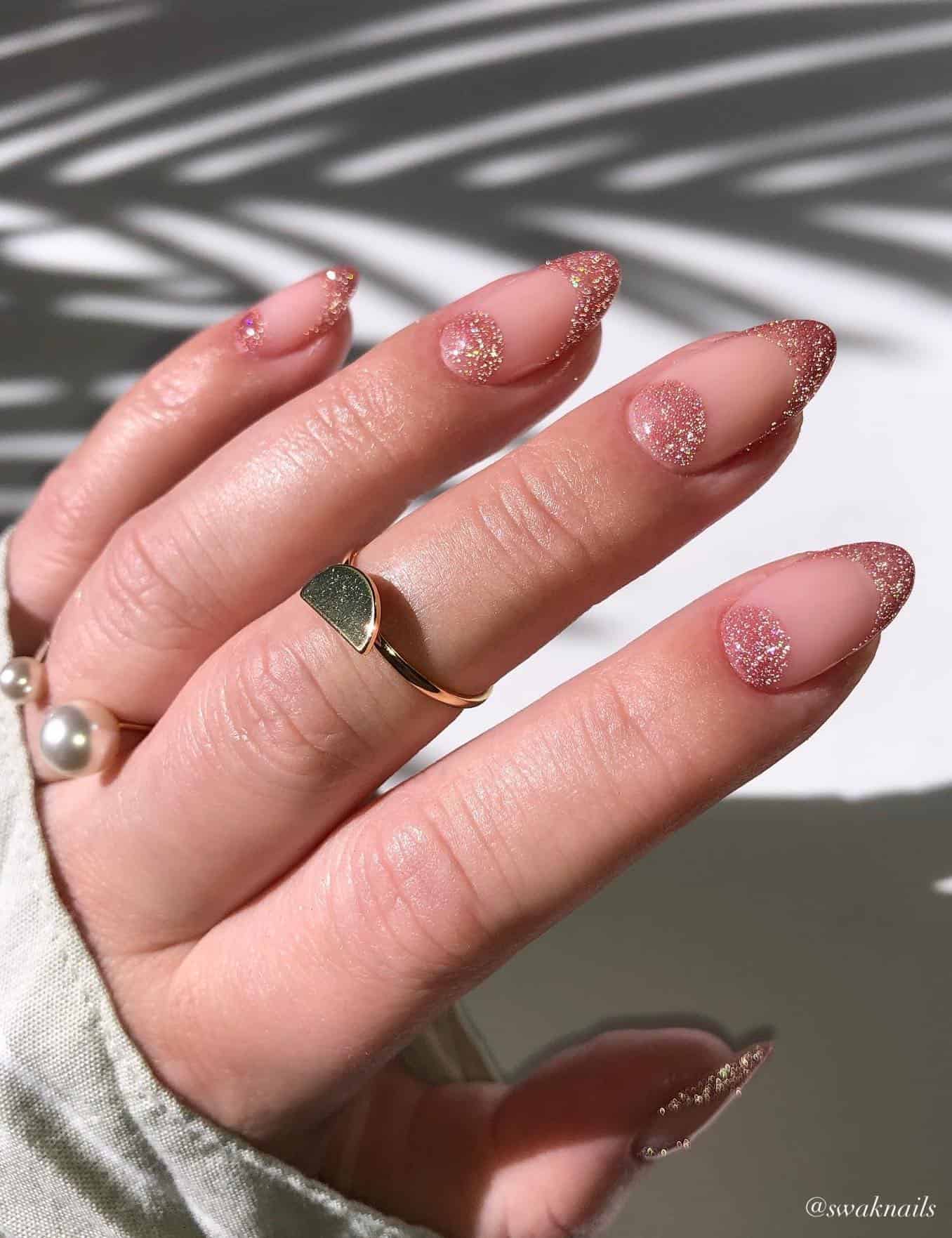 A hand with nude almond nails featuring French tips and half moon accents in a shimmering pink polish
