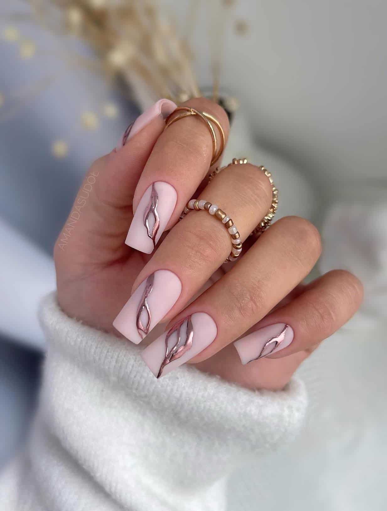 A hand with long square nails painted a matte pastel pink with metallic rose gold accents