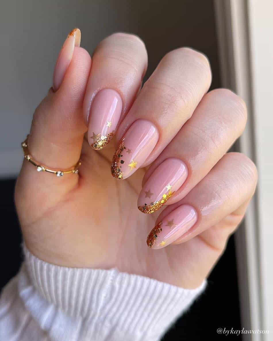 A hand with medium nude pink almond nails with asymmetrical gold glitter French tips
