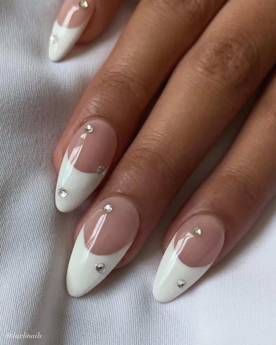 A hand with medium nude almond nails with white French tips and crystal accents