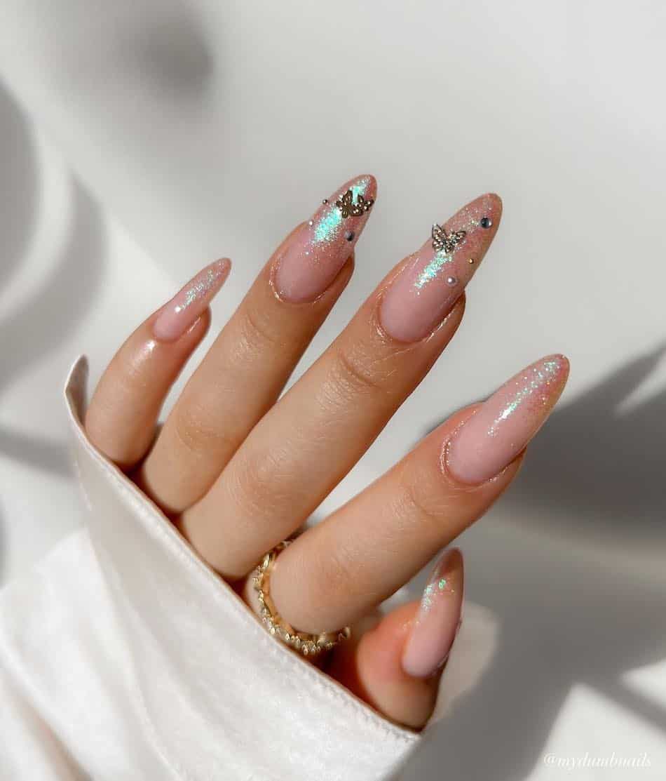 A hand with long nude almond nails with shimmering tips and butterfly charm accents