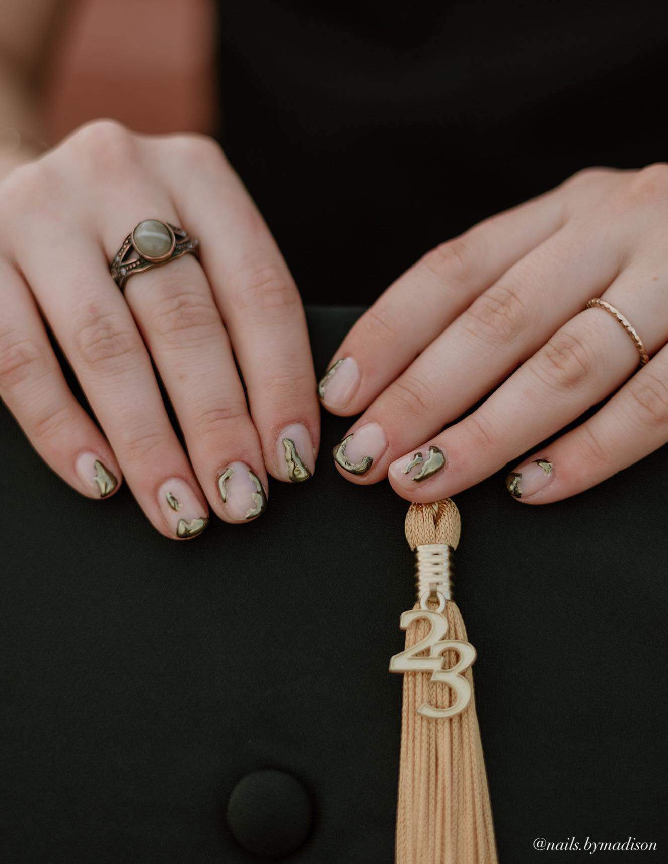 An image of hands holding a 2023 graduation cap with short nude nails with abstract gold accents