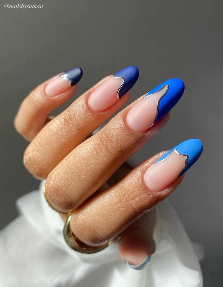 A hand with matte nude almond nails featuring gradient blue French tips with silver borders