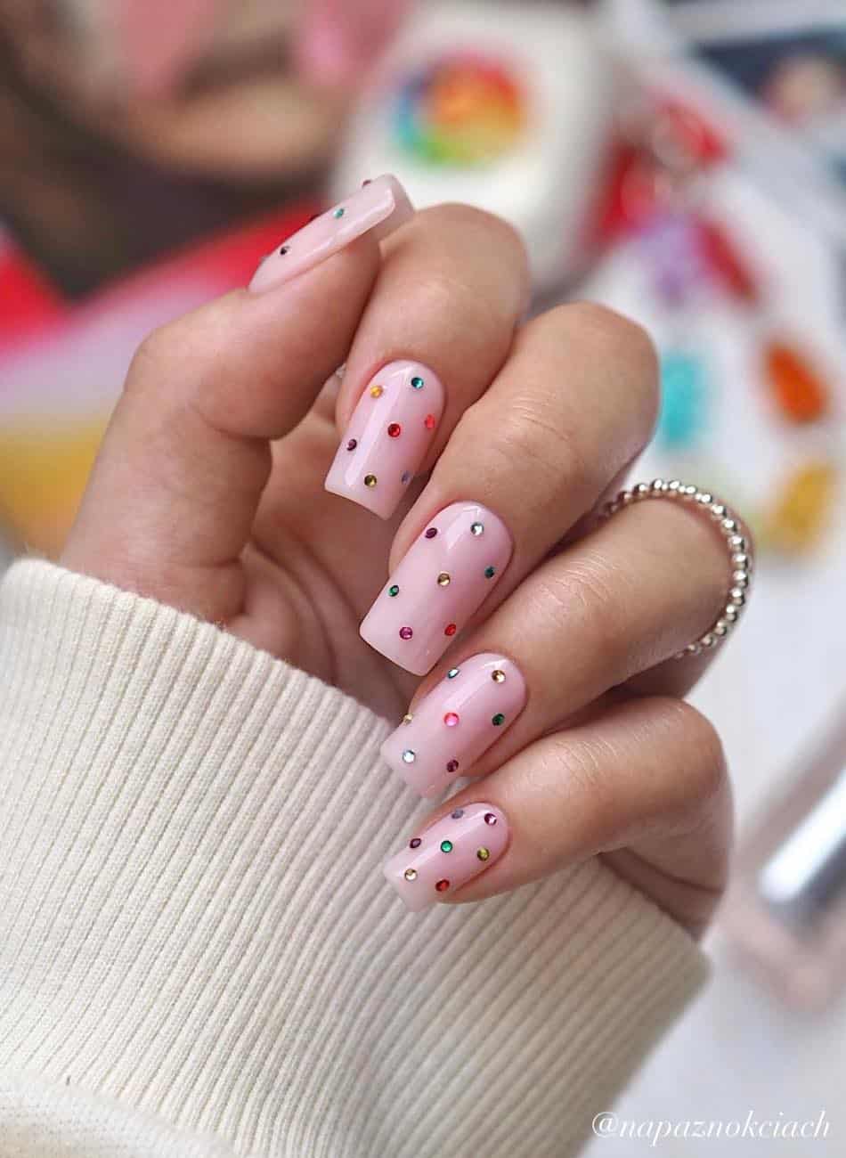 A hand with long nude pink square nails accented with colorful crystals