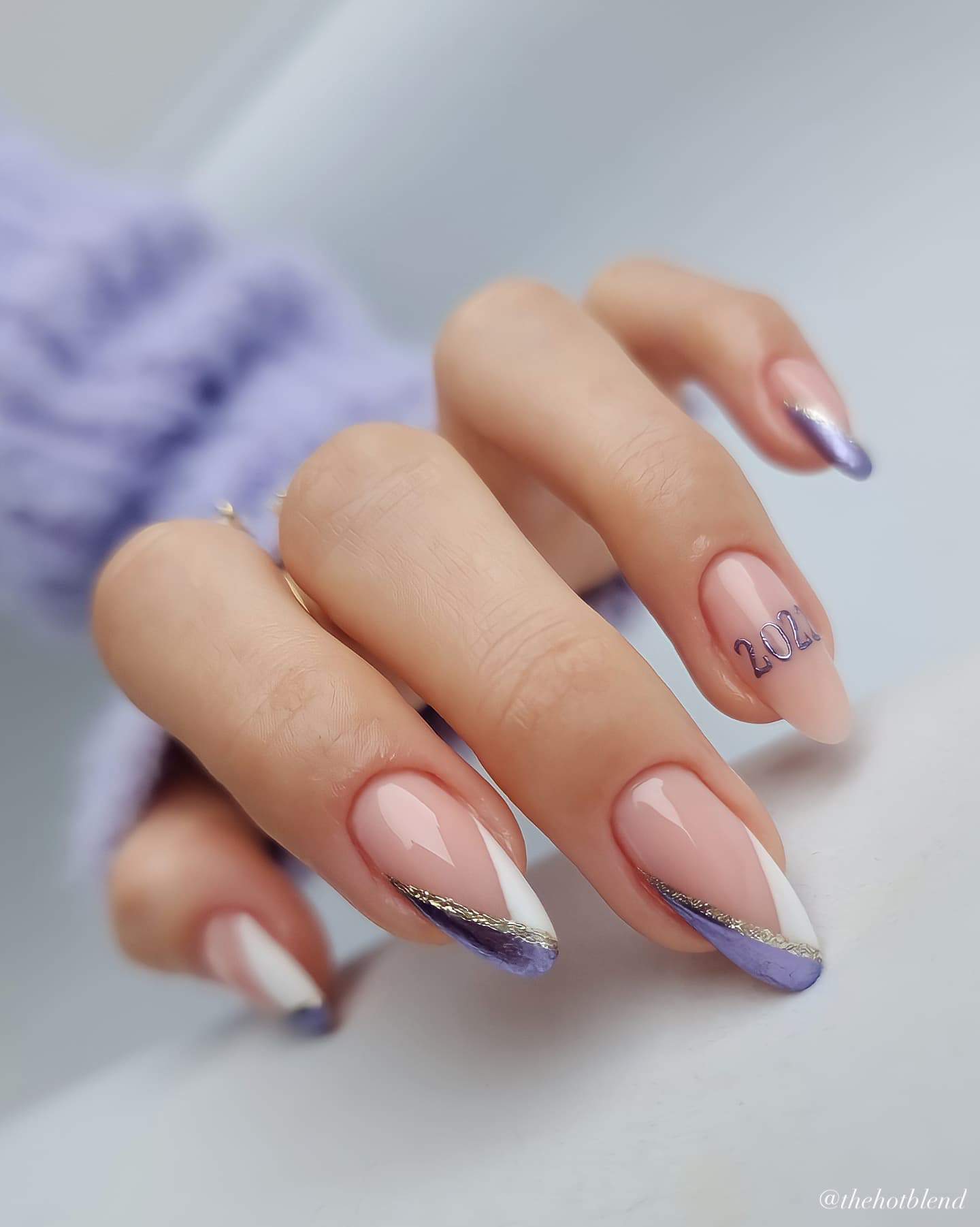 A hand with medium nude almond nails painted with silver, white, and purple French tips and an accent nail with a graduation year painted in polish