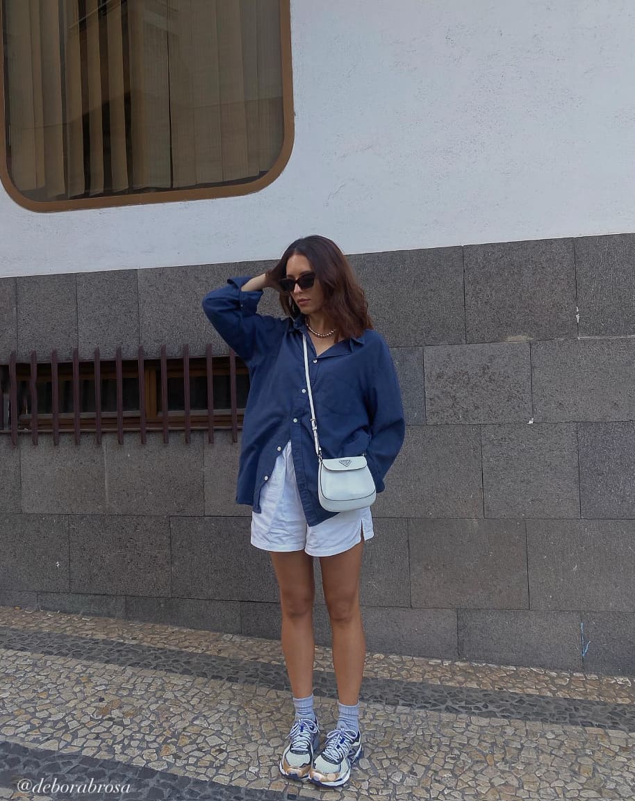A woman wearing white linen shorts with an oversized blue button-up, sneakers, and a pastel handbag