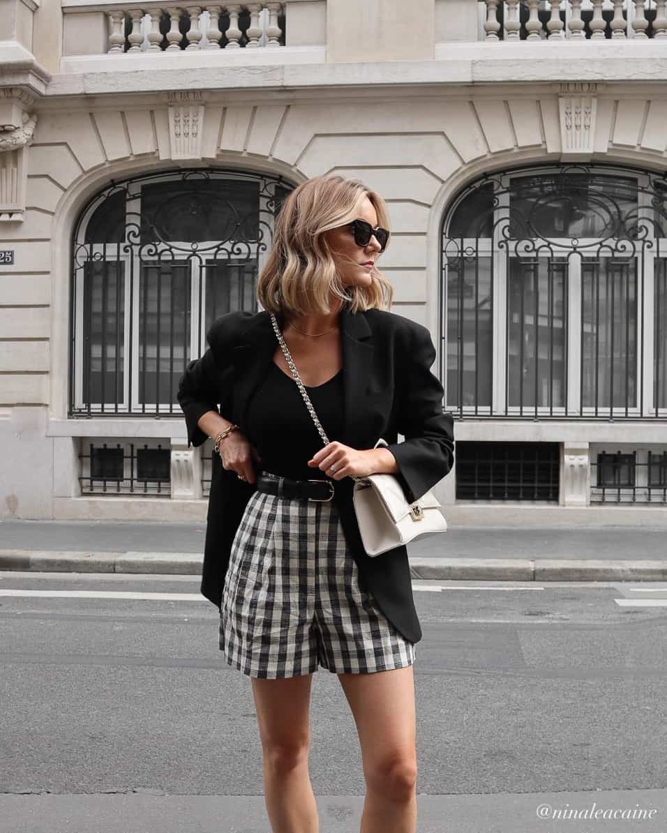 A woman wearing black and white checkered linen shorts with a black top, a black blazer, and a white handbag