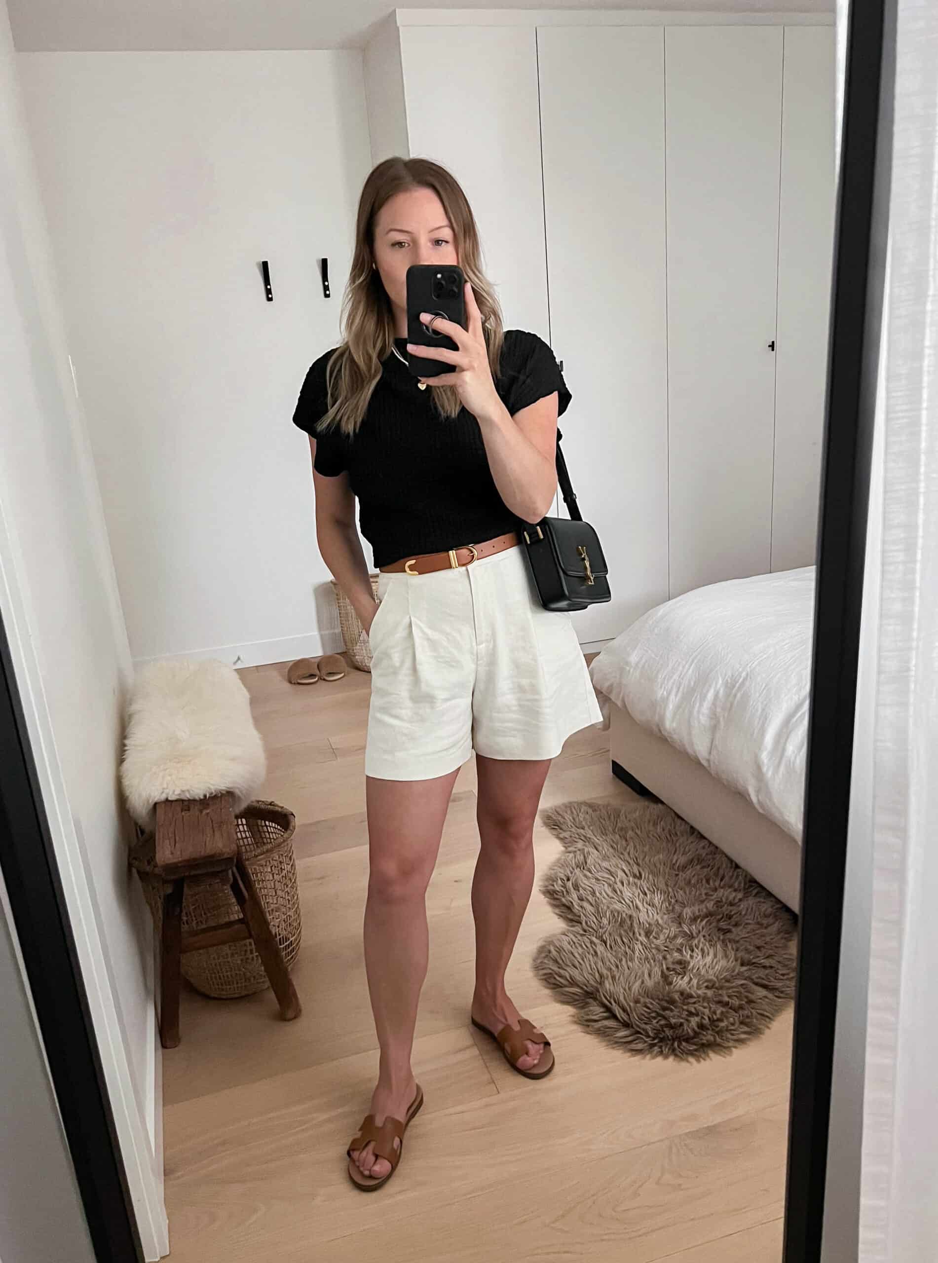 A woman wearing white linen shorts with a black top, tan sandals, and a black handbag