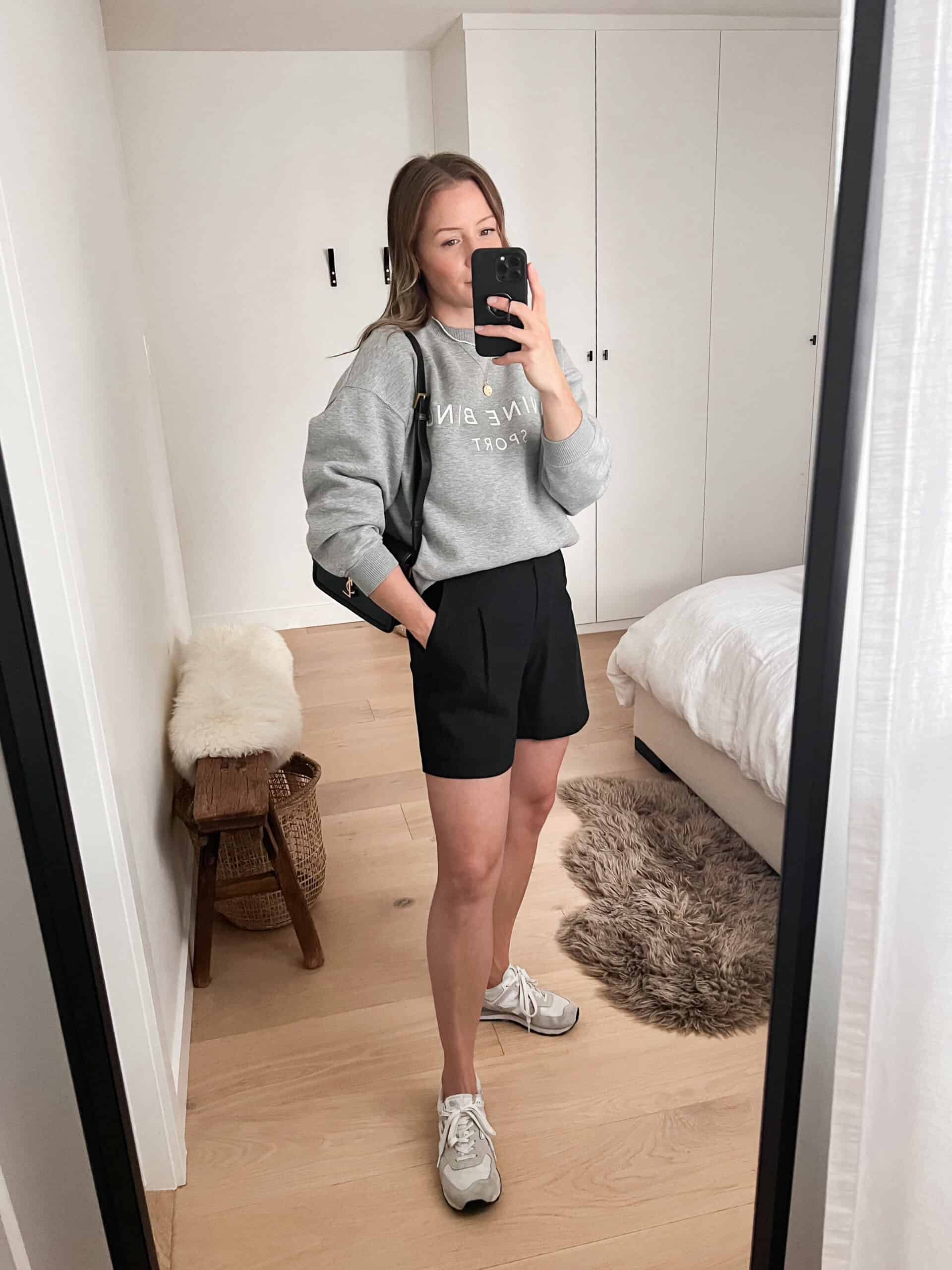 A woman wearing black linen shorts with a grey sweatshirt, trainers, and a black handbag