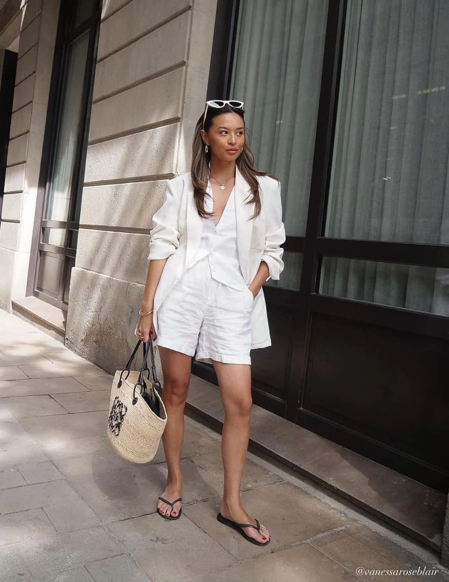 A woman wearing white linen shorts with a matching white linen vest, a white blazer, black flip flops, and a woven raffia tote