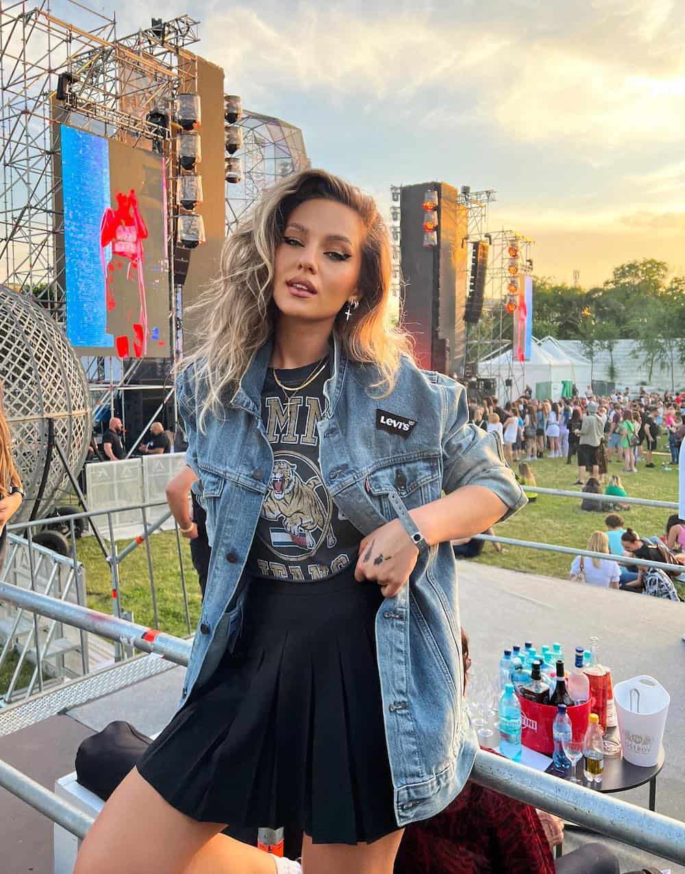 A woman at a concert wearing a black tennis skirt, a black graphic tee, and an oversized denim jacket