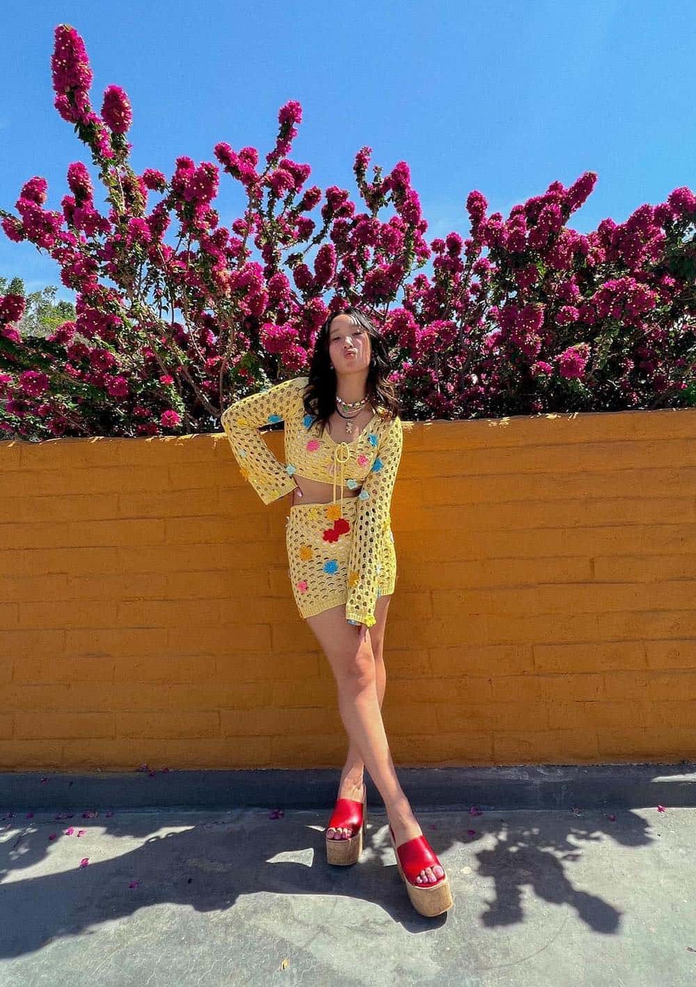 A woman wearing a yellow crochet set featuring a mini skirt and a long-sleeve cropped top with crocheted flowers and red platform sandals