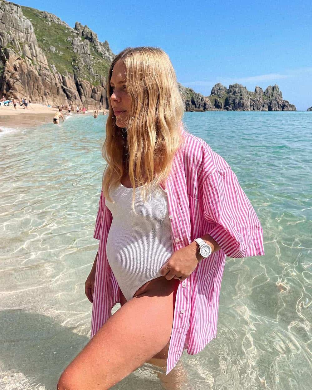 Pregnant woman wearing a white swimsuit with a striped pink button-up shirt over top