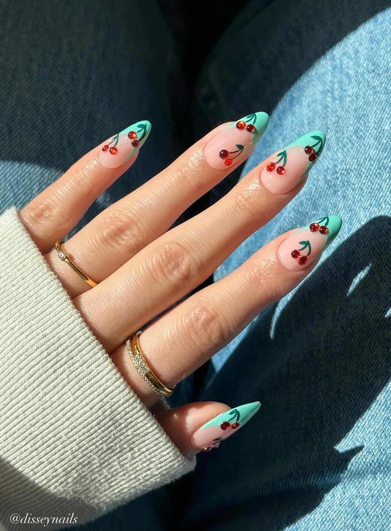 A hand with medium almond nails featuring mint green French tips and cherry nail art featuring red gem accents