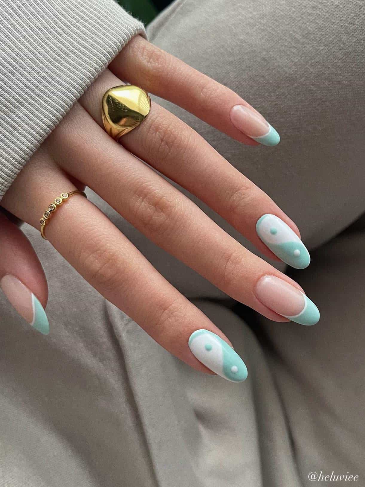 A hand with medium almond nails painted a glossy nude with mint green French tips with white outlines and two accent nails featuring white and mint green yin and yang nail art