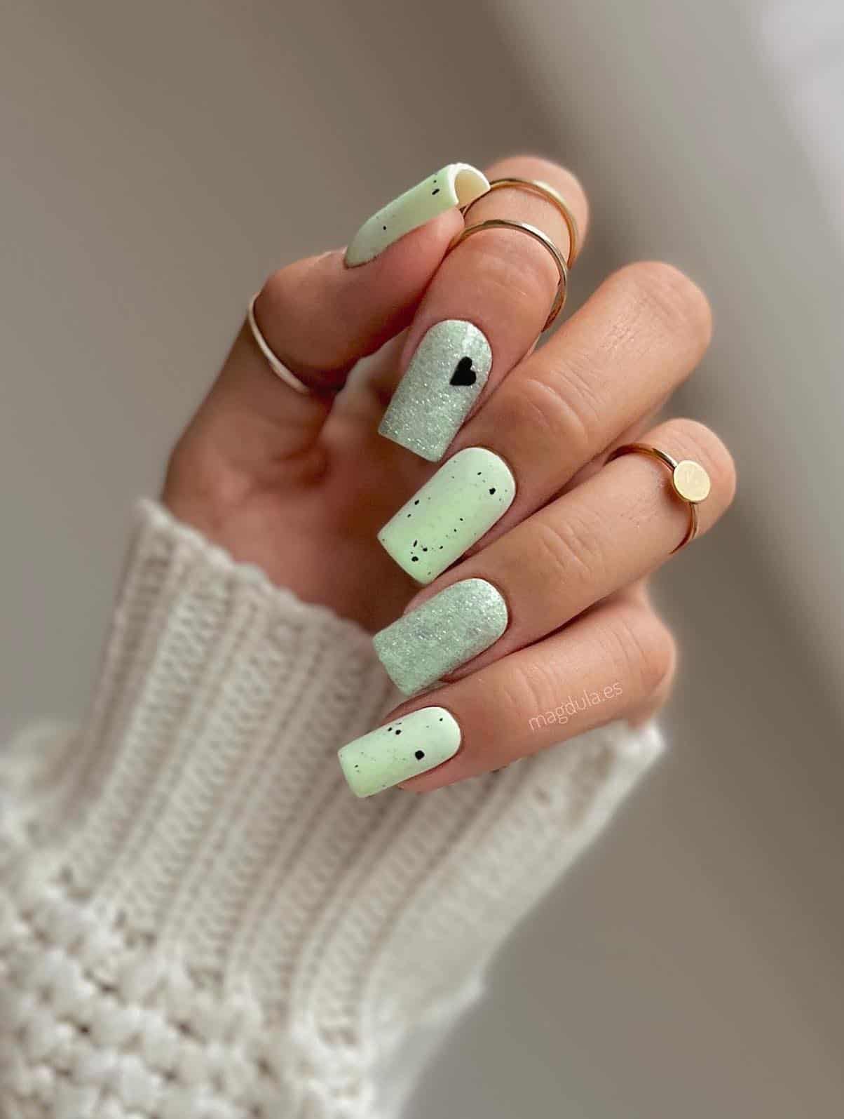 A hand with long square nails painted a mint green and speckled with black s=polish and two accent nails with a glitter topcoat and black heart detail