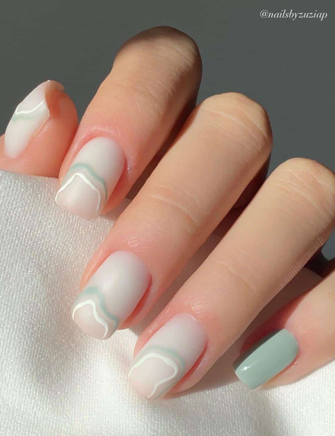 A hand with short matte white square nails with white and mint green abstract lines and one accent nail with glossy mint green polish