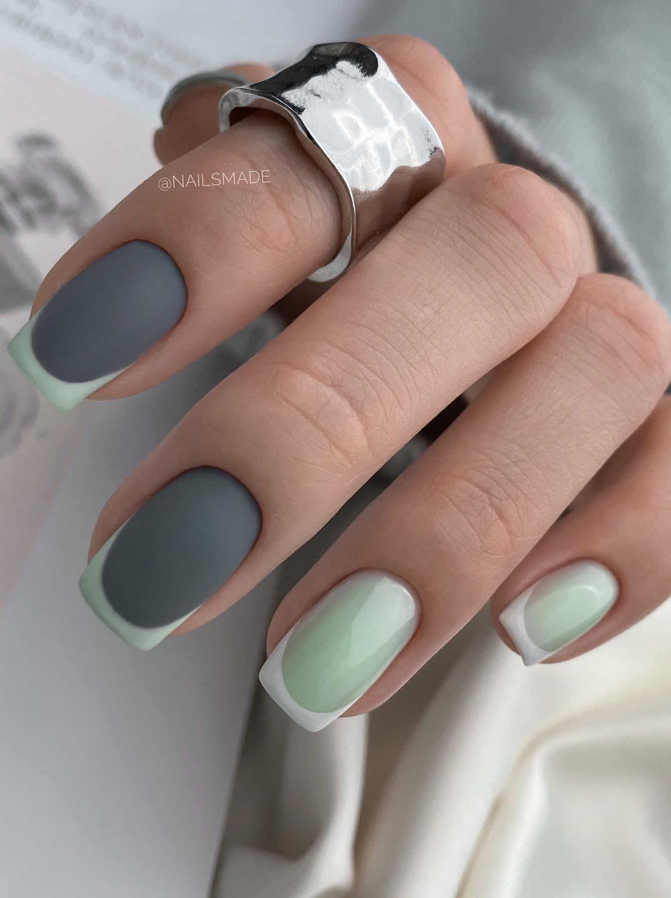 A hand with short square nails, half painted a glossy mint green with white French tips, and the other nails are a dark matte grey with mint green French tips
