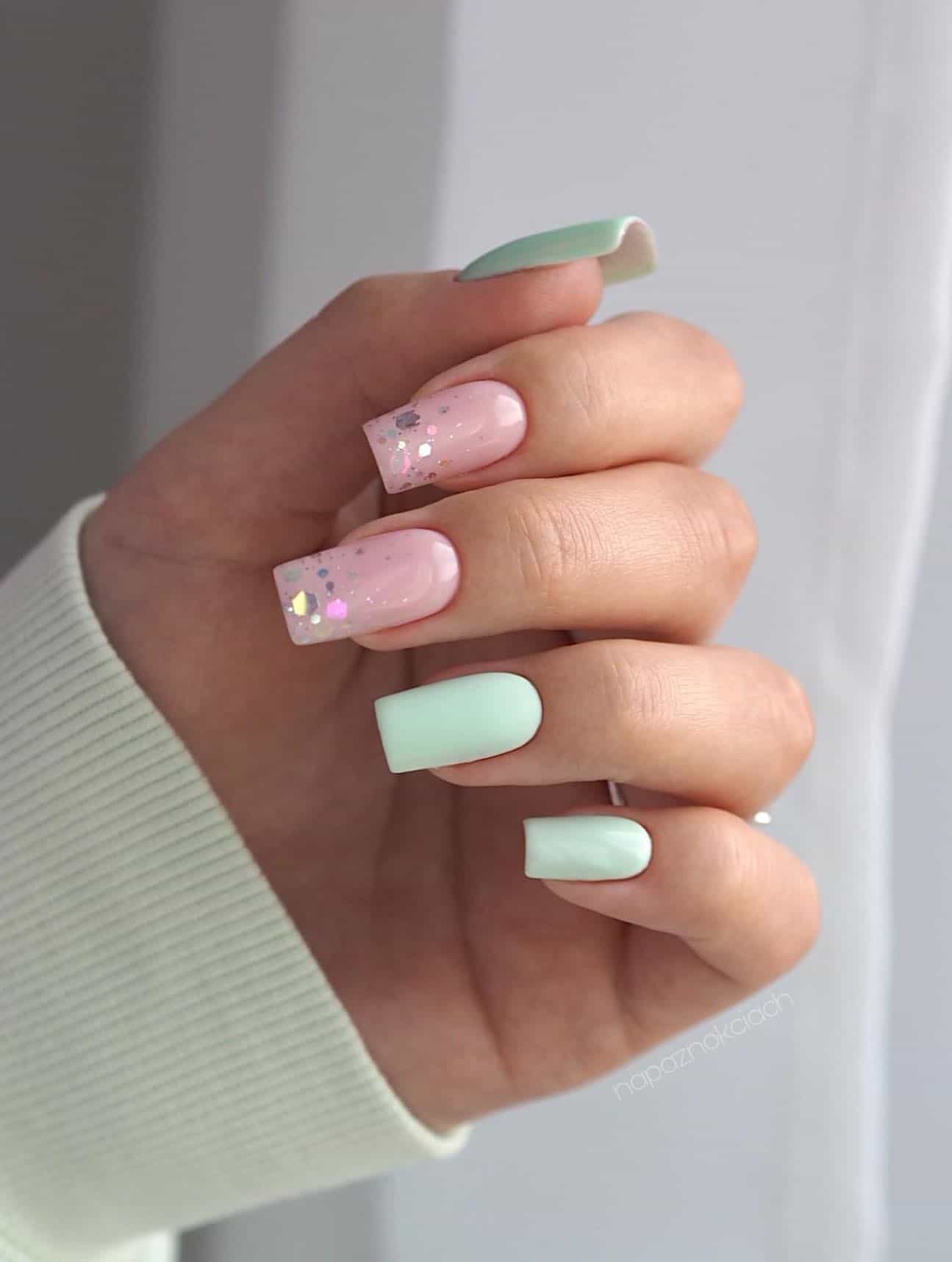 A hand with medium square shaped nails painted mint green with two nude pink accent nails with chunky silver glitter tips