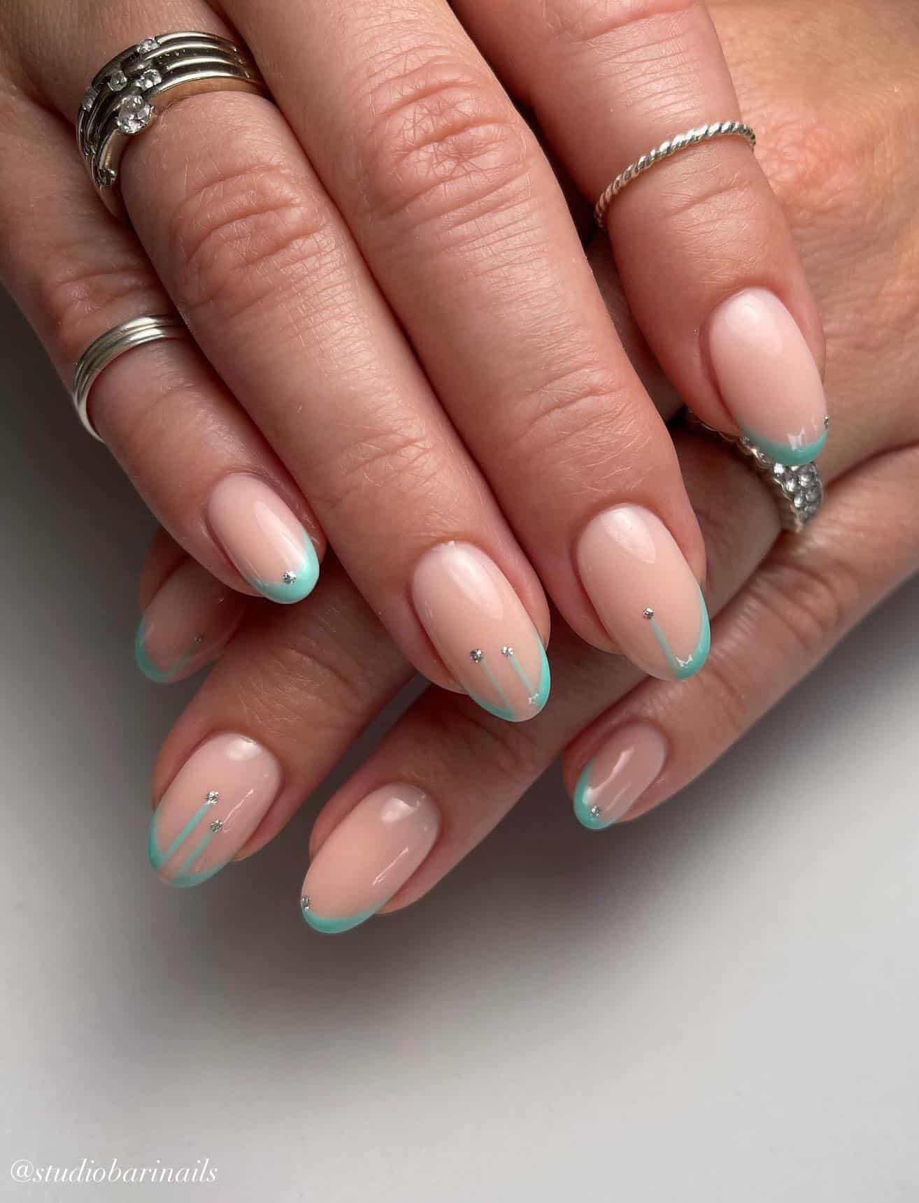 A hand with nude almond nails with mint green French tips painted with a dripping effect and silver gem accents