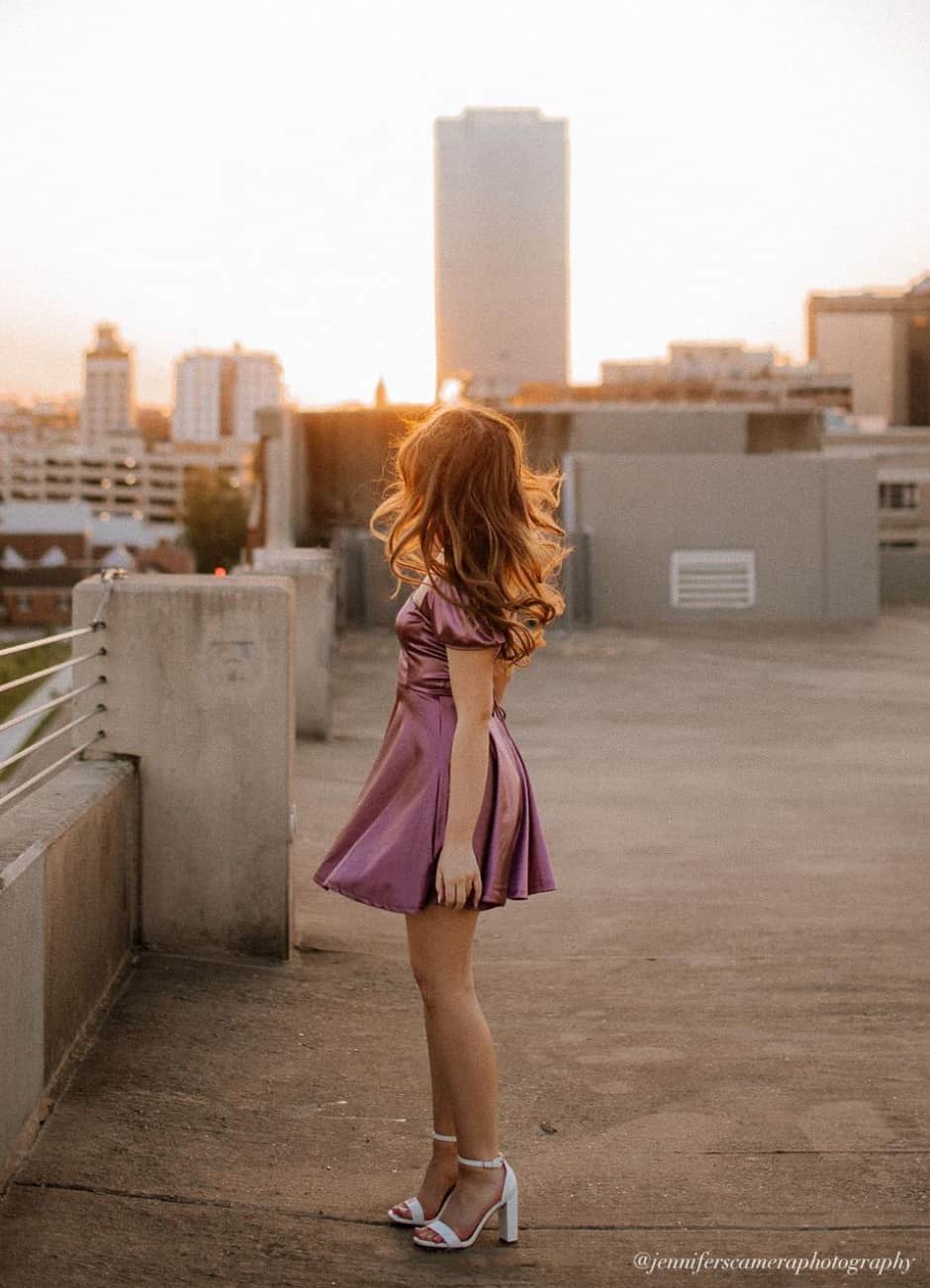 Young woman wearing a purple satin mini dress with white heeled sandals for senior pictures