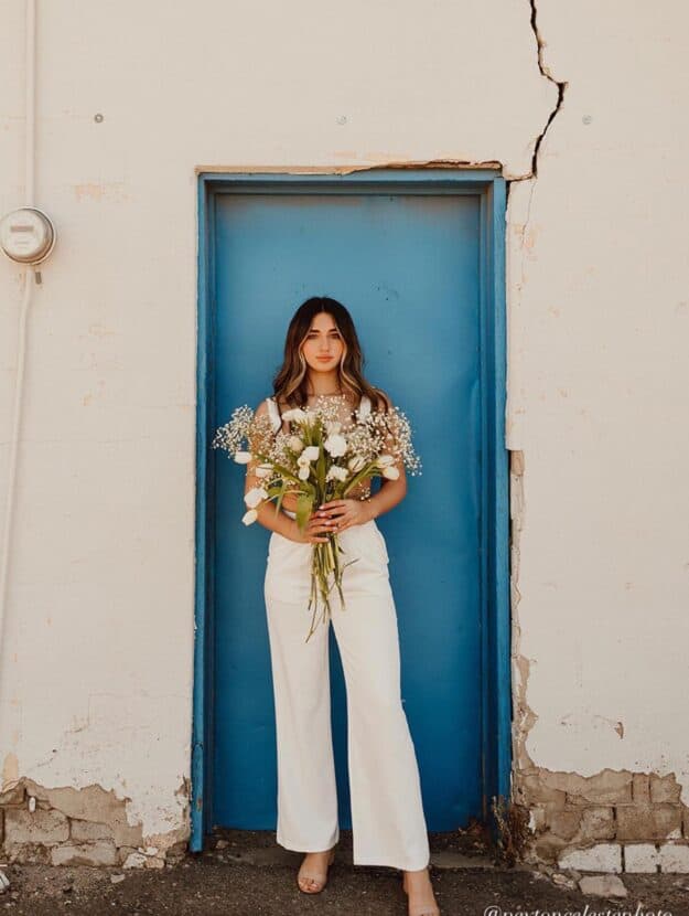 Young woman standing in front of a blue door with a bouquet of flowers wearing a white matching pant set for senior pictures