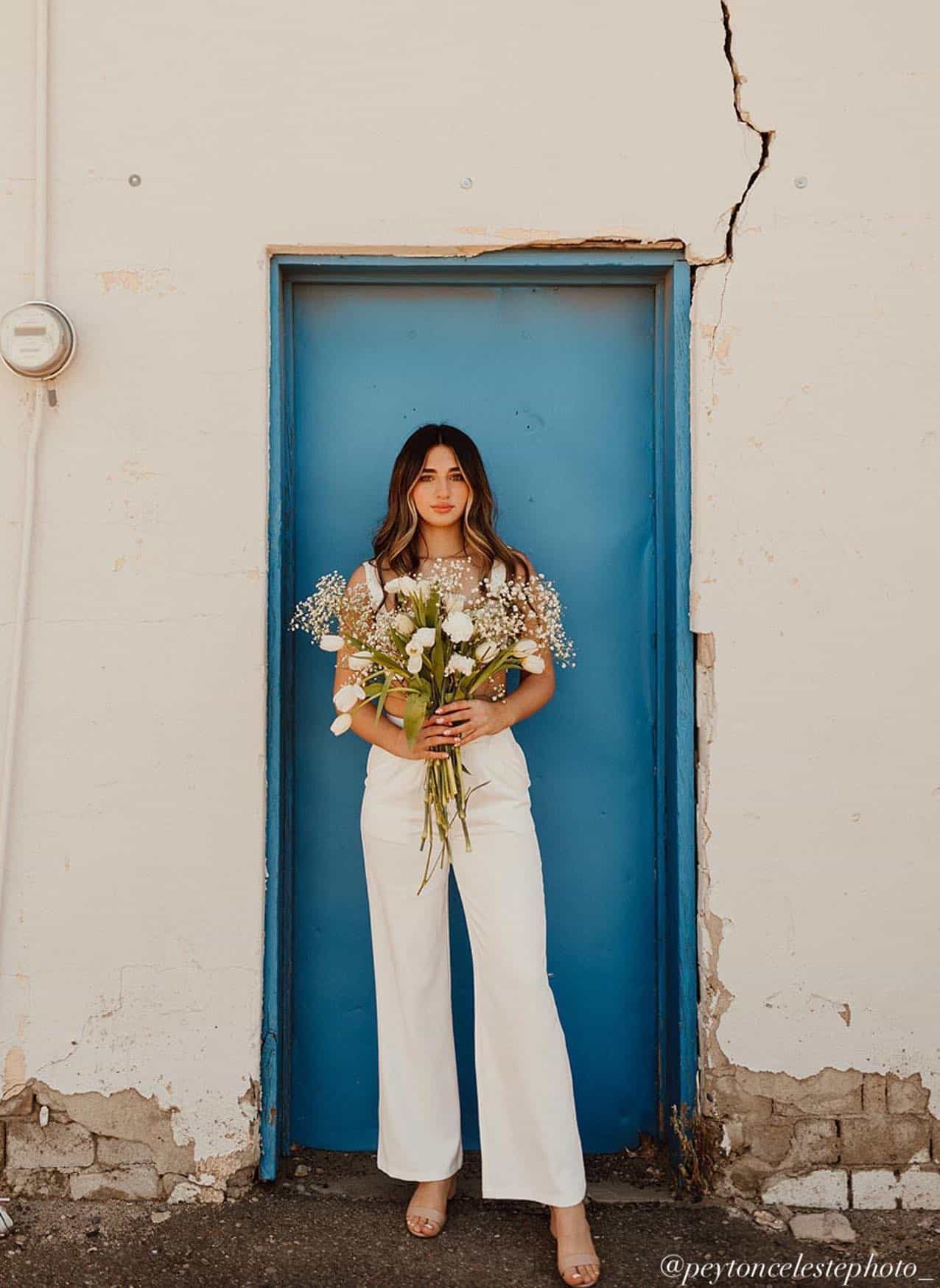 Young woman standing in front of a blue door with a bouquet of flowers wearing a white matching pant set for senior pictures