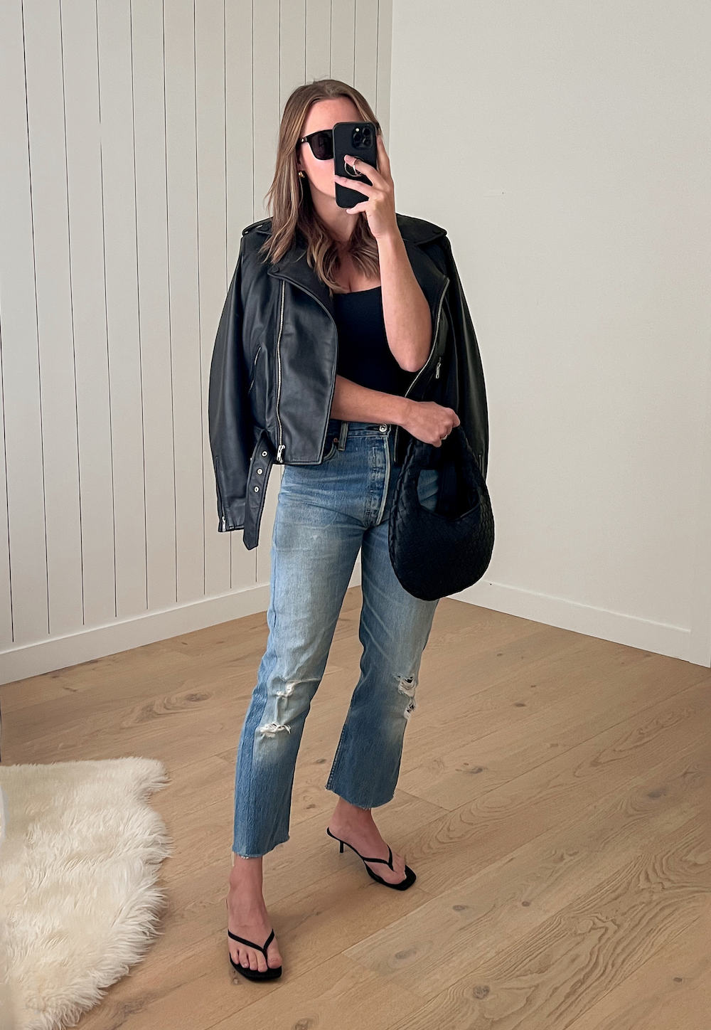 Woman wearing distressed straight leg jeans, a black tank top and a black leather jacket with black heeled sandals.