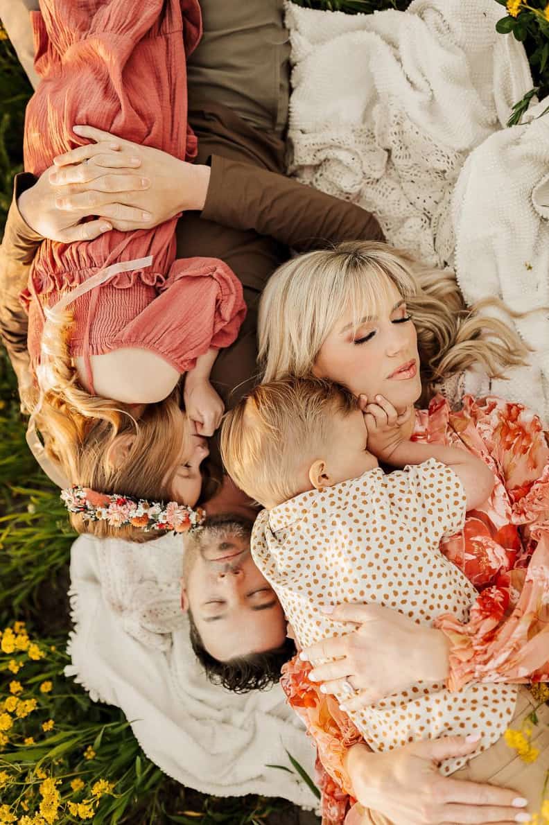 An image of a summer family photo with a family of four laying on a blanket in a field and holding each other, wearing red and pink toned clothing and neutrals