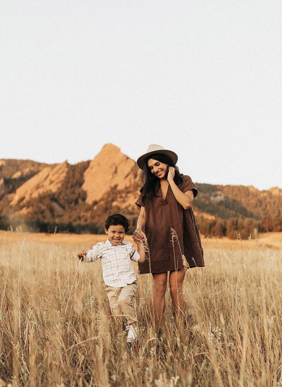 An image of a mother and her son in a field for summer family photos, the mother wearing a short brown dress and fedora while the son wears a plaid button-up and beige khakis