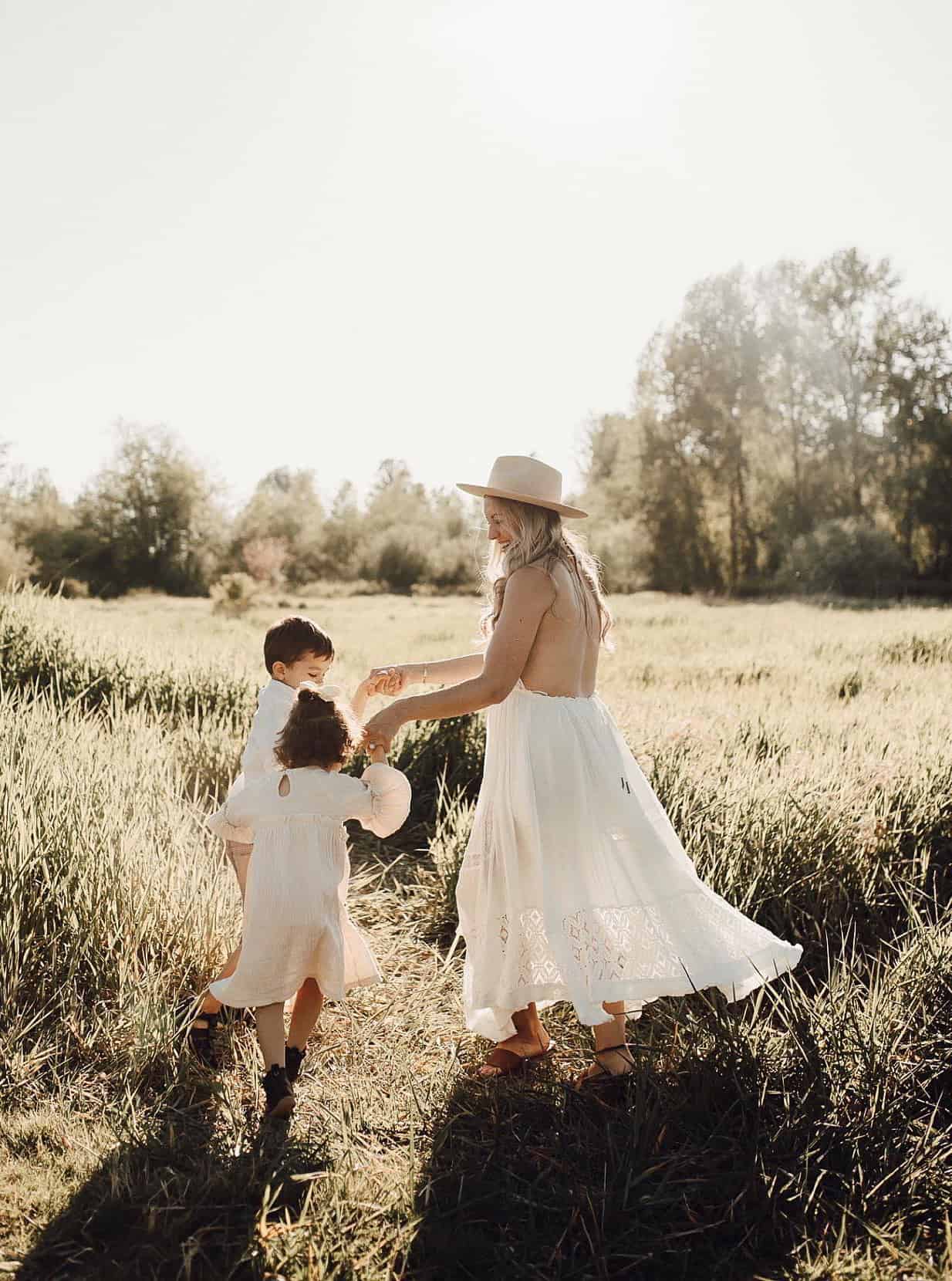 An image of a summer family portrait with A mother wearing a white summer dress and fedora dancing in a field with her children