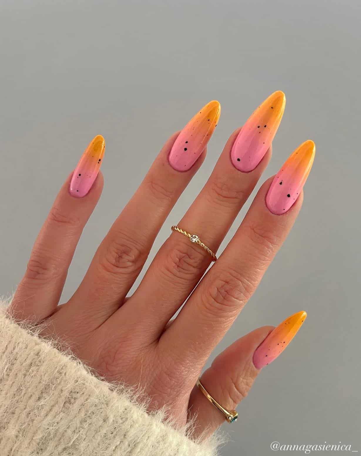 A hand with long almond nails featuring an orange and pink ombre with black speckles