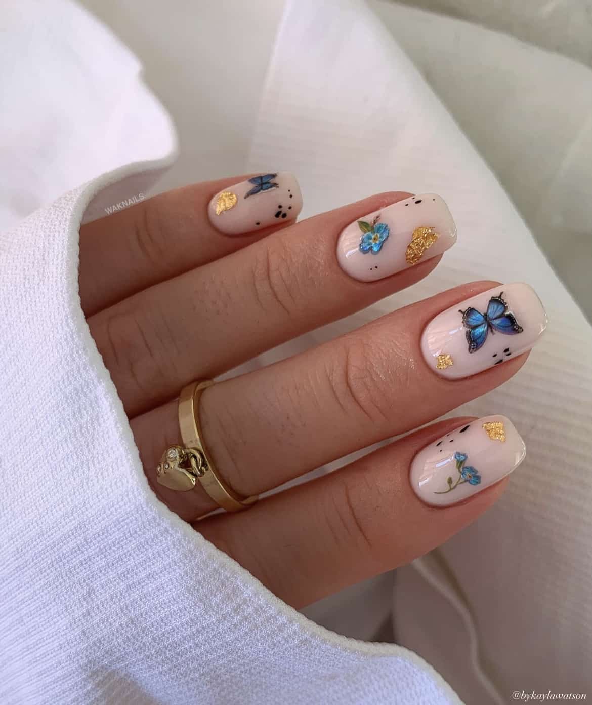A hand with short squoval nails painted a milky white color and accented with gold flakes, blue butterflies, and blue flowers