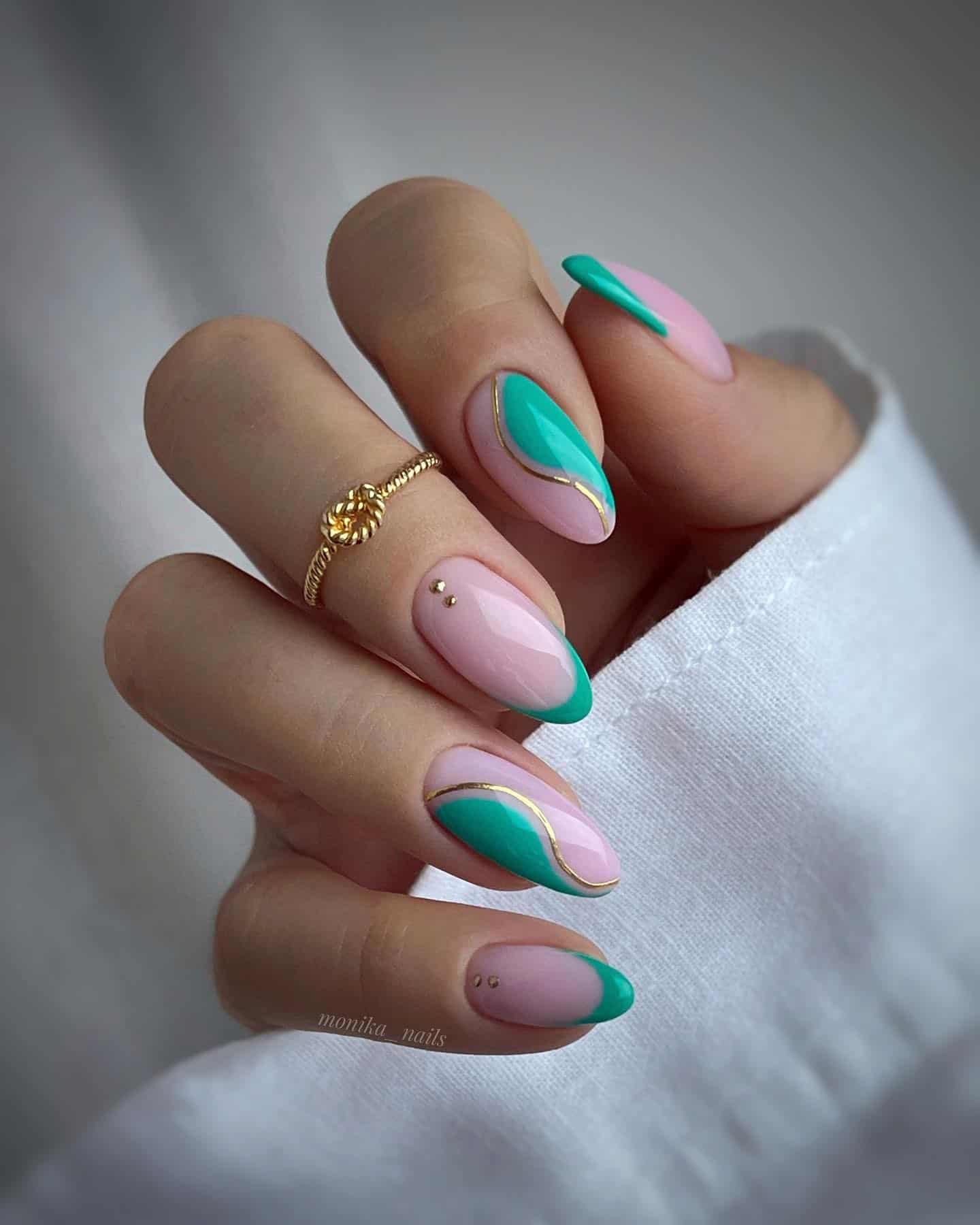 A hand with medium almond nails painted a light pink with green wave accents, green French tips, and gold line details