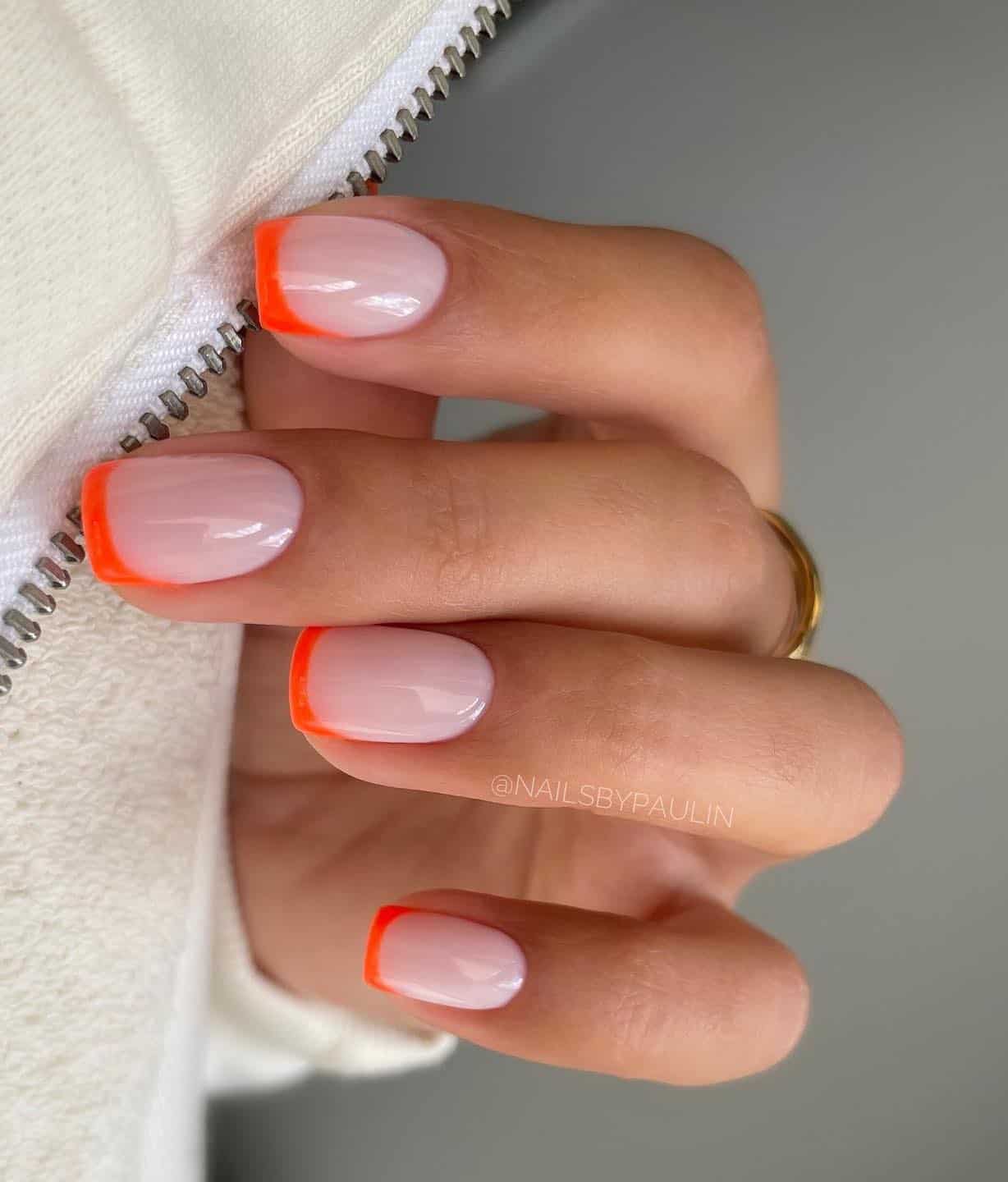 A hand with short square nails painted a pinkish nude tone with bright orange French tips