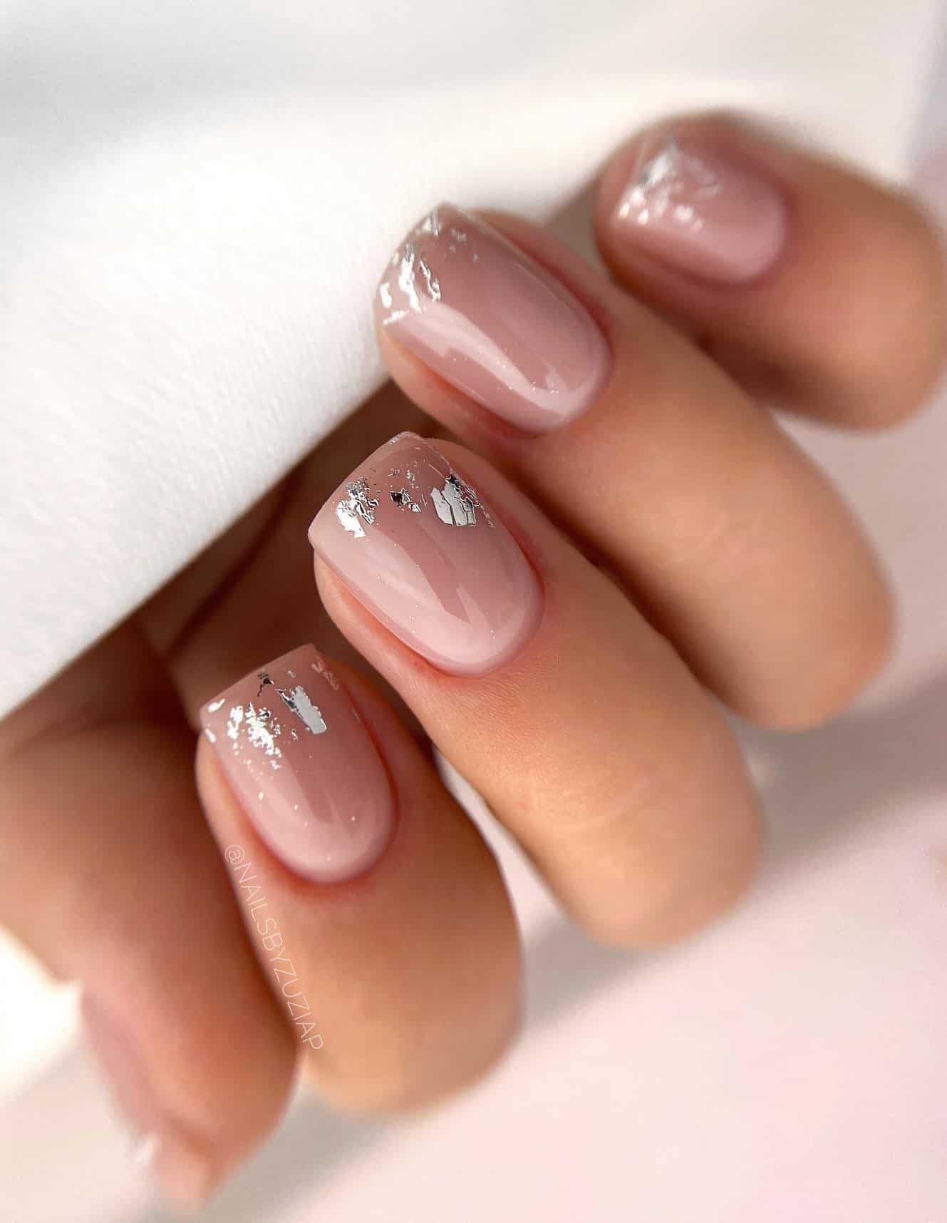A hand with short square nails painted a nude color with silver flakes along the tips