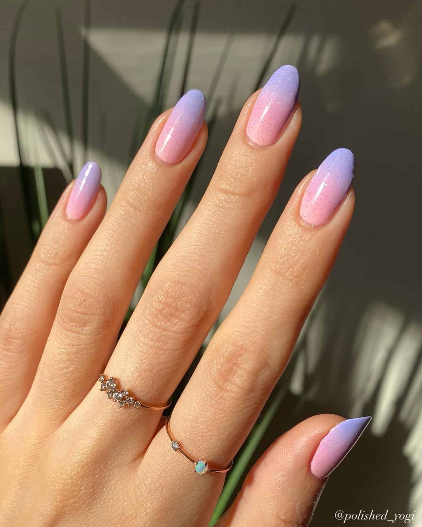 A hand with medium almond nails painted with a pink and purple ombre design and a glittering topcoat