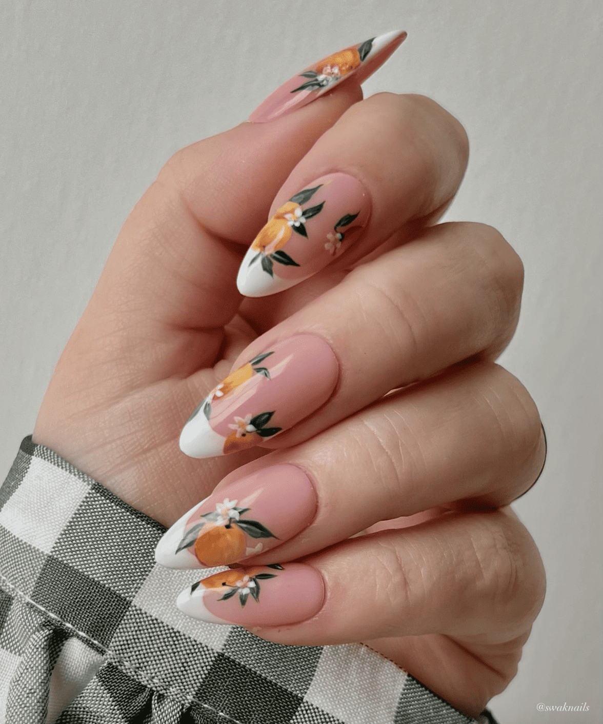 A hand with long almond nails featuring white French tips and nail art of oranges with leaves and flowers