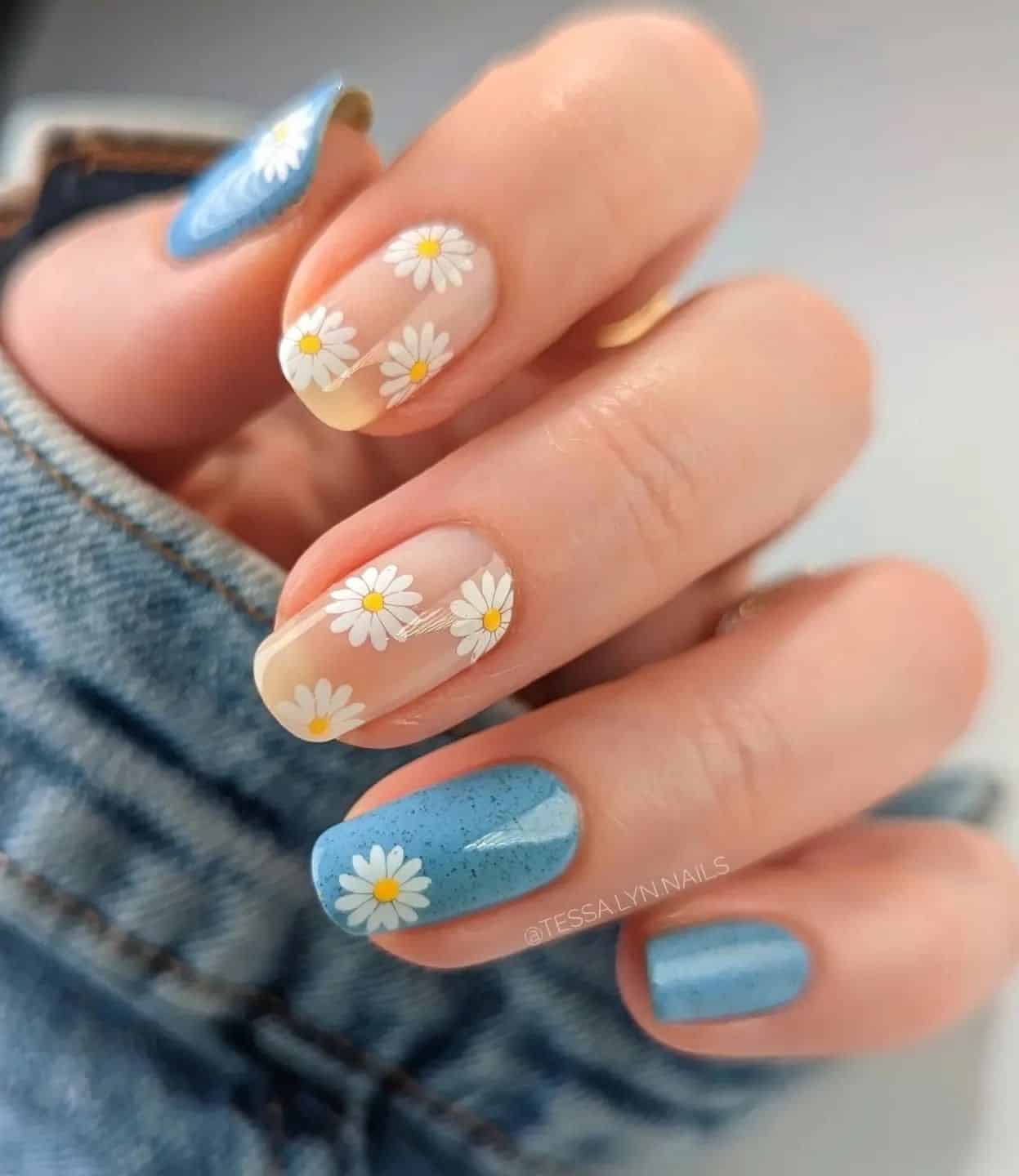 Una mano con uñas cortas y curvas que presenta una mezcla de uñas nude y azules con un arte de uñas en forma de margarita blanca.