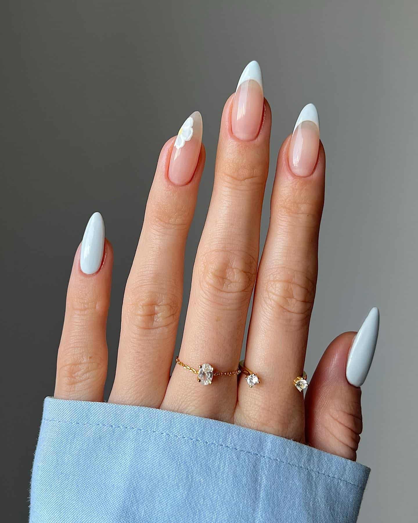 A hand with medium almond nails painted in classic French tips with one accent nail featuring a small white flower along the side