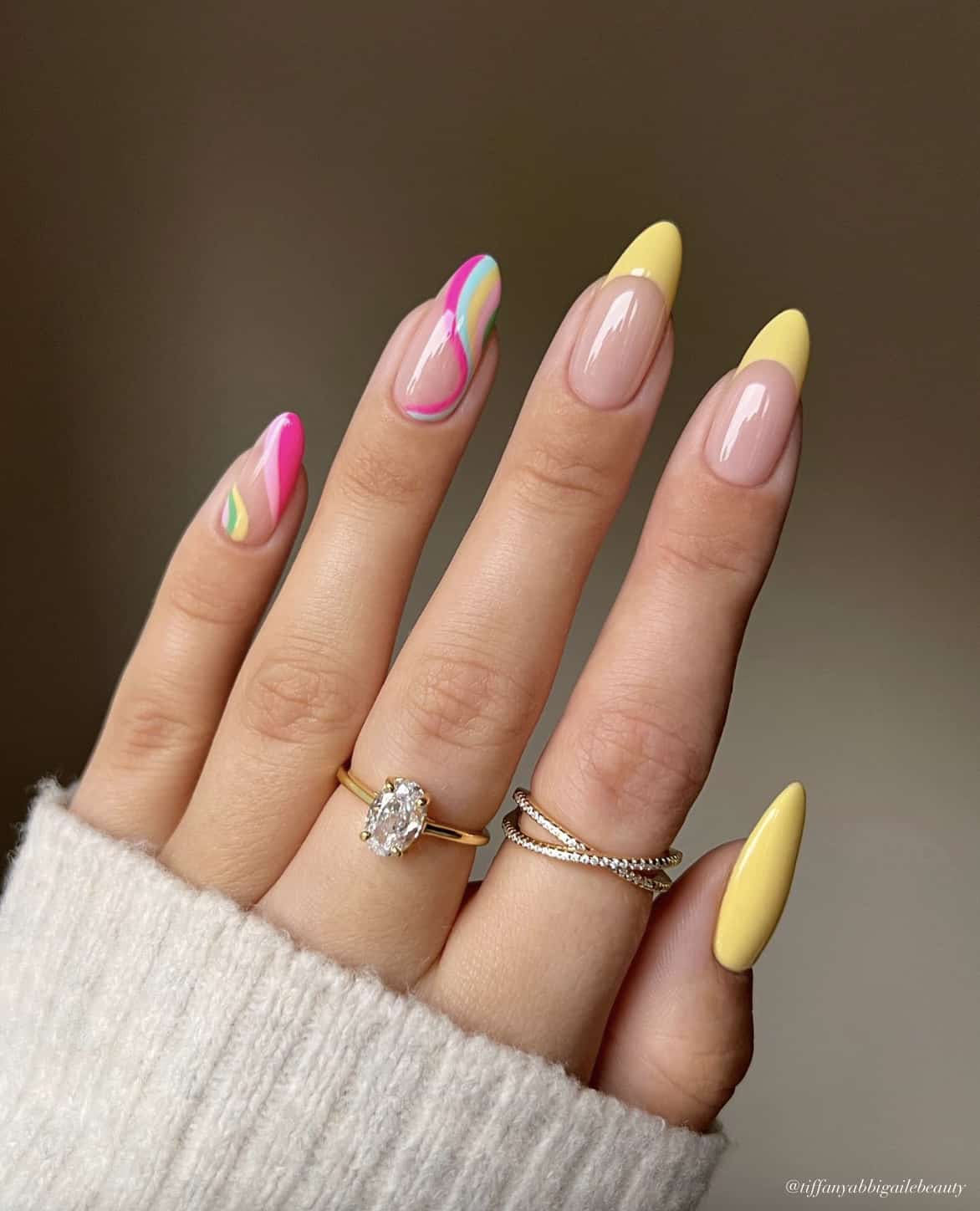 A hand with medium almond nails painted with various summer nail designs, including yellow French tips, a solid yellow nail, and yellow, blue, and pink swirled nails