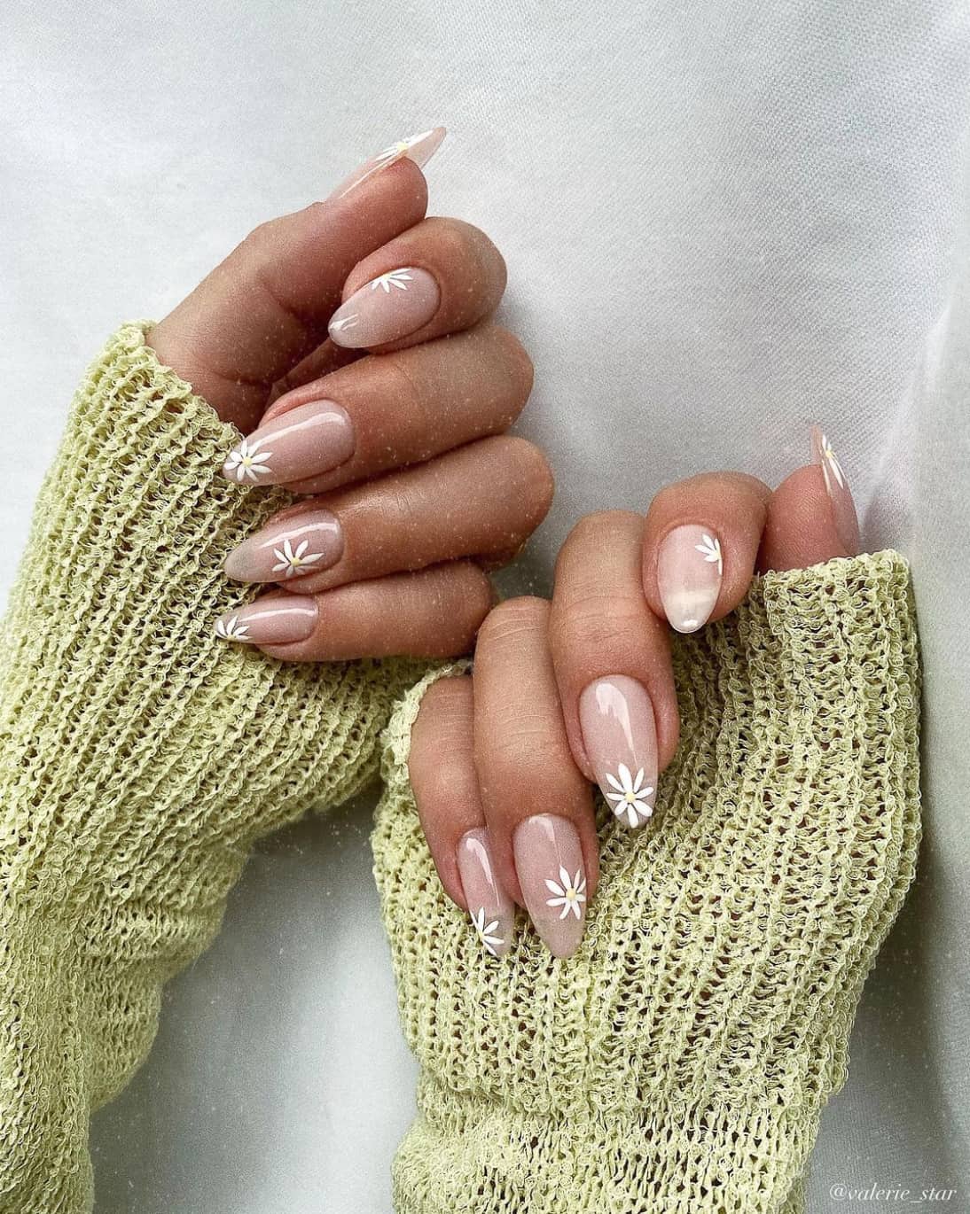 A hand with medium almond nails painted a nude tone and accented with white flowers