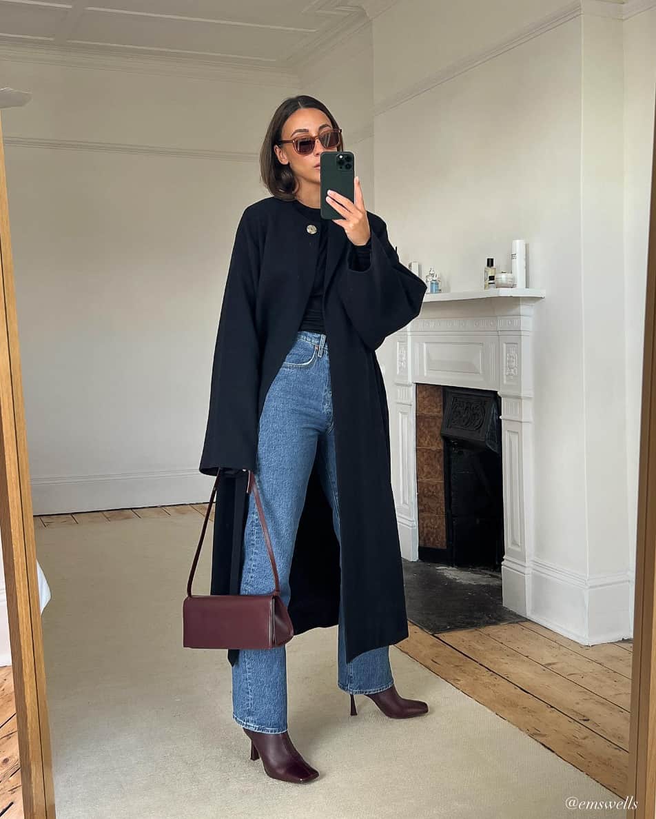 woman wearing a long navy coat with straight leg jeans in medium blue wash and burgundy ankle boots with a burgundy bag