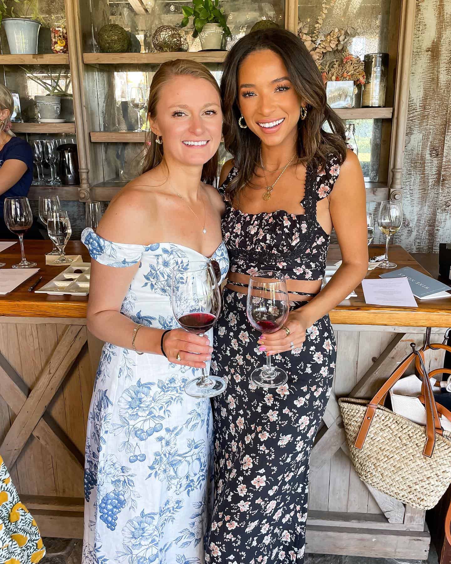 two women wearing floral dresses holding glasses of red wine at a winery
